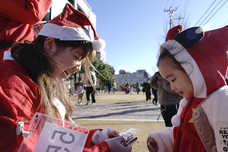 サンタが女の子のビンゴカードを受け取って、自身の番号を開けている様子。強風で髪がゆれているが、二人はほほ笑んでいる。