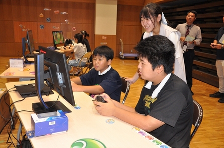 中学生くらいの男の子二人が対戦している。後ろから中学生から高校生くらいの女の子が微笑みながら見守っている。