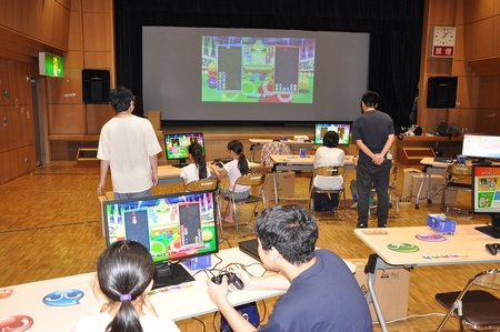 会場の様子。ゲーム機とモニターが各机にいくつか準備されている。一番前にスクリーンに投影されたぷよぷよゲームの画面が映っている。