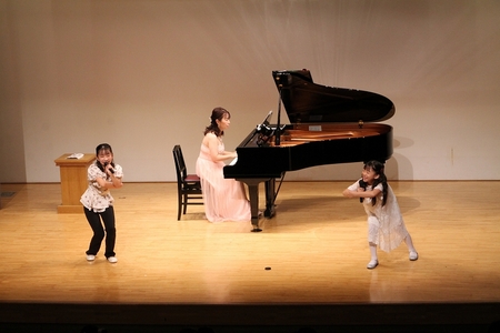 佐々木朋子さんがピアノを演奏し娘2人が歌いながら手遊びを実演している